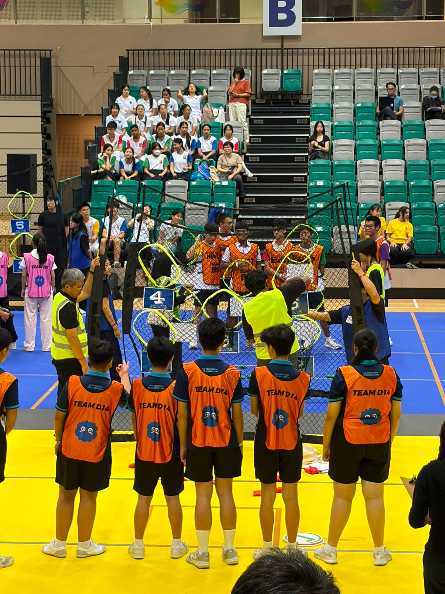 A Hai Sing Catholic School student in PE uniform, wearing an apron, blonde wig, and lei, acting out a roleplay or dramatisation scene next to a small table labeled 'Aunty Loh STOVE' during an English Language and Literature lesson